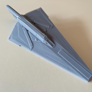 Op de afbeelding: Lichtgrijs model van een futuristisch ruimteschip, met een centraal lichaam en schuine vleugels. Het model heeft gedetailleerde paneellijnen en een gestroomlijnd ontwerp. Het lijkt op een miniatuur ruimteschip of vliegtuig, mogelijk een verzamelobject of display-item.