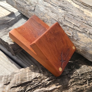 May include: A handcrafted wooden phone stand in a rich reddish-brown hue. The stand features a curved design to cradle a phone, with a decorative emblem and two small, round accents. The stand is made of wood and is set on a weathered wooden surface.