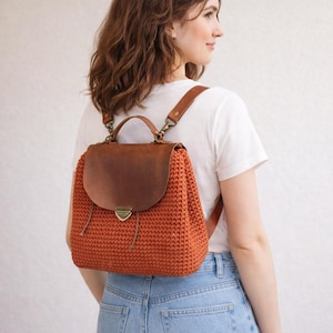 Może przedstawiać: Pomarańczowy plecak z szydełka z brązową skórzaną klapą i paskami. Torba ma metalowe zapięcie i górny uchwyt. Paski są regulowane. Plecak jest noszony na ramionach.