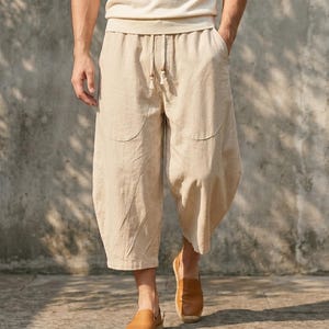 May include: Beige linen culottes with a drawstring waist and curved pocket details. The cropped pants are paired with a cream-colored top and brown leather loafers. The casual outfit is suitable for warm weather.