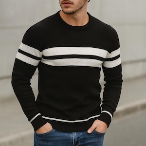 以下が含まれることがあります： 黒いクルーネックセーター。胸に2本の白い水平ストライプと、袖に1本の白いストライプがあります。セーターは、テクスチャードニット生地で作られています。モデルは青いジーンズを着用しています。