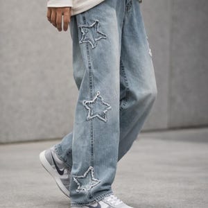 May include: Light blue denim jeans with star-shaped cutouts. The jeans have a relaxed fit and feature distressed details. The star cutouts are outlined with a lighter thread, adding a unique design element. The jeans are paired with grey and white trainers.