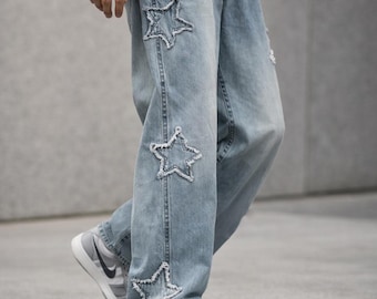 May include: Light blue denim jeans with star-shaped cutouts. The jeans have a relaxed fit and feature distressed details. The star cutouts are outlined with a lighter thread, adding a unique design element. The jeans are paired with gray and white sneakers.