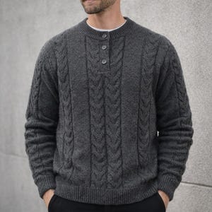 Może przedstawiać: Szary sweter z warkoczowym splotem z zapięciem na guziki. Sweter ma okrągły dekolt i długie rękawy. Projekt obejmuje pionowe wzory warkoczowe i prążkowany dół. Modelka ma na sobie białą koszulkę.