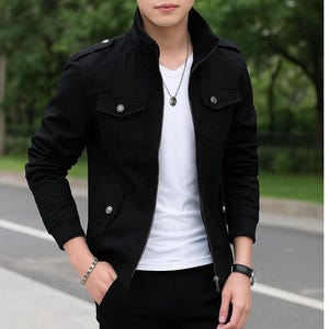 Könnte beinhalten: Schwarze Jacke mit Stehkragen, zwei Brusttaschen und Reißverschluss. Die Jacke hat Knöpfe an den Schultern und Taschen. Das Model trägt ein weißes T-Shirt darunter.