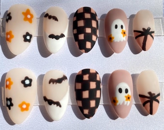 May include: A set of ten oval-shaped false nails. Designs include orange and black flowers, black bats, black and peach checkered patterns, a white ghost with orange flowers, and a peach nail with a black bow.