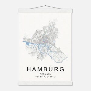 Könnte beinhalten: Ein weißes Wandbild mit einer detaillierten Karte von Hamburg, Deutschland, mit blauen Wasserwegen und einem roten Marker. Der Name der Stadt und die Koordinaten sind unten in Schwarz gedruckt. Die Karte wird von Holzstäben gehalten.