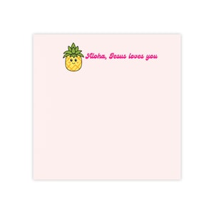 Könnte beinhalten: Ein quadratischer, hellrosa Notizblock mit einer kleinen, gelben Cartoon-Ananas mit grünem Blattwerk und einem lächelnden Gesicht. Neben der Ananas ist der Text "Aloha, Jesus loves you" in einer rosa Schreibschrift gedruckt.
