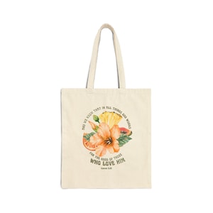 May include: A beige canvas tote bag featuring a central graphic of a large orange hibiscus flower, a pineapple slice, and citrus fruit segments. The text reads: AND WE KNOW THAT IN ALL THINGS GOD WORKS FOR THE GOOD OF THOSE WHO LOVE HIM ROMANS 8:28.