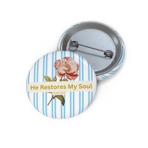 May include: Two round pinback buttons. One button features a pink and cream-colored flower with green leaves on a light blue and white striped background. The text "He Restores My Soul" is displayed on a cream-colored banner. The other button is silver.