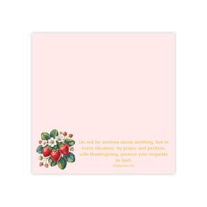 May include: A light pink square notepad with a floral strawberry illustration in the lower left corner. The illustration features red strawberries, white flowers, and green leaves. Gold text reads, "Do not be anxious about anything..." and a Bible verse.