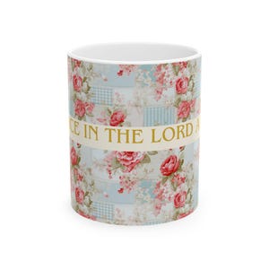 May include: A white ceramic mug with a floral design featuring red roses and a light blue patchwork pattern. The mug has a gold band with the text "Grace in the Lord".