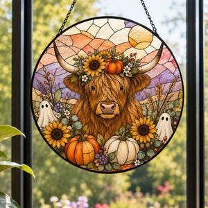 May include: A circular stained glass suncatcher featuring a brown Highland cow with long horns, adorned with a sunflower crown. The scene includes orange and white pumpkins, small white ghost figures, and wheat stalks against a sunset sky.
