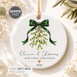 May include: A round white ceramic ornament featuring a green mistletoe illustration tied with a dark green ribbon. The text reads Olivia & Thomas, OUR FIRST CHRISTMAS, and 2026, with a small gold heart icon centered above the year and a hanging twine loop.