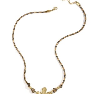 Könnte beinhalten: Halskette mit goldfarbenem Blumenanhänger und gemustertem Kordel. Die Halskette hat einen goldfarbenen Blumenanhänger in der Mitte und eine Kordel mit schwarzen, braunen und goldenen Akzenten. Die Halskette hat einen Karabinerverschluss und eine verstellbare Kette.