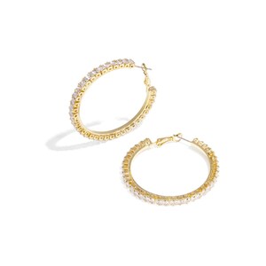 Könnte beinhalten: Zwei goldfarbene Creolen. Jeder Ohrring ist mit kleinen, klaren Edelsteinen besetzt. Die Ohrringe sind vor einem weißen Hintergrund platziert, mit einem Schatten.