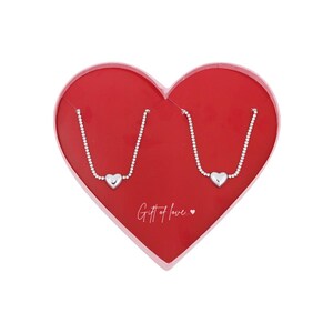 Könnte beinhalten: Zwei silberne Halsketten mit Herzanhängern werden in einer roten herzförmigen Schachtel präsentiert. Auf der Schachtel steht "Gift of love". Die Halsketten befinden sich auf weißem Hintergrund.