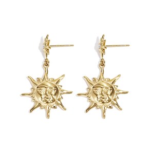 Könnte beinhalten: Ein Paar goldfarbene Ohrringe mit einem Sonnengesicht-Design. Jeder Ohrring hat einen sternförmigen Stecker und einen hängenden Sonnenanhänger mit detailliertem Gesicht und Strahlen. Auf weißem Hintergrund.