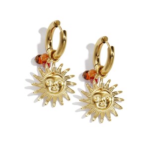 Könnte beinhalten: Ein Paar goldfarbene Creolen. Jeder Ohrring hat einen Sonnenanhänger mit einem lächelnden Gesicht und einer Ansammlung von orangefarbenen Perlen. Die Ohrringe sind zum Tragen am Ohr bestimmt.