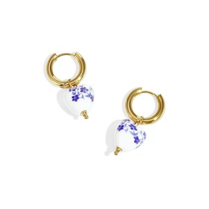 Könnte beinhalten: Ein Paar goldfarbene Creolen mit herzförmigen Anhängern. Die weißen Keramikherzen sind mit einem blauen Blumenmuster verziert. Die Ohrringe werden vor einem weißen Hintergrund präsentiert.