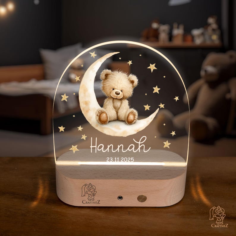 A wooden nightstand with a lighted display featuring a teddy bear sitting on a crescent moon and surrounded by stars. The display has a personalized name and date, indicating it is a gift for Hannah.