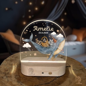 a wooden stand with a clear glass front, displaying a picture of a whale and stars. The name "Amelie" is written above the whale, and the date "23.11.2025" is below the whale.