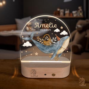 A wooden nightstand with a lighted glass display featuring a blue whale and stars, along with the name "Amelie" and the date "23.11.2025".