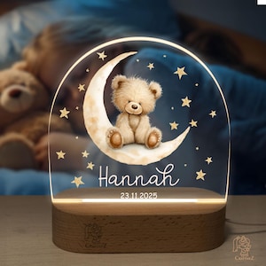 A teddy bear sitting on a crescent moon, surrounded by stars, is displayed on a wooden stand with the name "Hannah" and the date "23.11.2025" written on it.