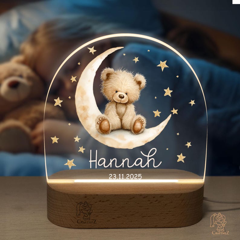 A teddy bear sitting on a crescent moon, surrounded by stars, is displayed on a wooden stand with the name "Hannah" and the date "23.11.2025" written on it.