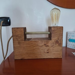 Puede incluir: Lámpara de mesa rústica de madera con una bombilla Edison de color ámbar cálido. La lámpara tiene una base rectangular de madera, una barra metálica horizontal y un cable de alimentación texturizado. La lámpara está sobre una mesa de madera.