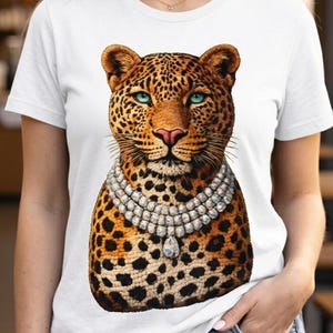 May include: White t-shirt featuring a detailed illustration of a leopard's face with blue eyes and a diamond necklace. The leopard has a spotted coat. The shirt is a classic crew neck style.