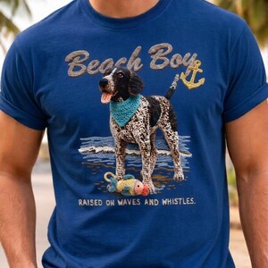 May include: Navy blue t-shirt with a graphic of a dog wearing a bandana, standing on a beach with a fish. Text reads "Beach Boy" and "Raised on Waves and Whistles."