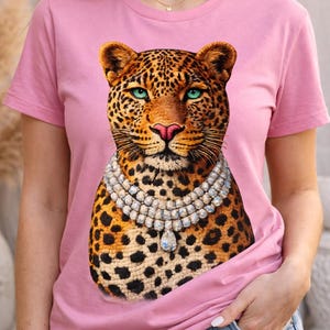 May include: Pink t-shirt featuring a leopard graphic with a jeweled necklace. The leopard has a spotted coat, blue eyes, and a pink nose. The shirt is a solid pink color.