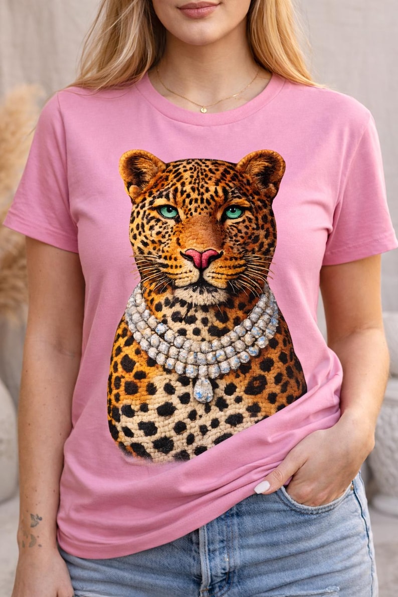 May include: Pink t-shirt featuring a leopard graphic with a jeweled necklace. The leopard has a spotted coat, blue eyes, and a pink nose. The shirt is a solid pink color.