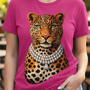 May include: A magenta t-shirt featuring a leopard design. The leopard has a spotted coat, blue eyes, and a diamond necklace. The shirt is short-sleeved and worn with blue jeans.