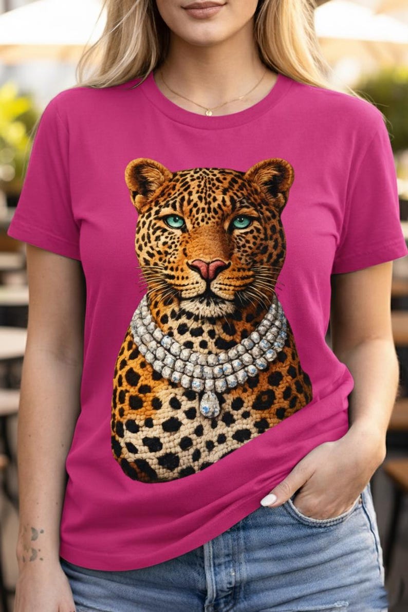 May include: A magenta t-shirt featuring a leopard design. The leopard has a spotted coat, blue eyes, and a diamond necklace. The shirt is short-sleeved and worn with blue jeans.