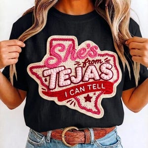 May include: Black t-shirt with a crochet Texas-shaped patch. The patch features the text "She's from TEJAS I CAN TELL" in pink and red. The shirt is paired with blue jeans and a brown belt.