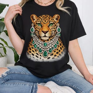 May include: Black t-shirt featuring a leopard illustration with a jeweled collar and earrings. The leopard has green eyes and a spotted coat. The jewelry is silver and emerald green. The shirt is a crew neck.