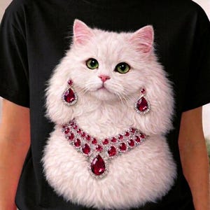 May include: Black t-shirt featuring a white cat with green eyes, pink nose, and ears. The cat is adorned with a red jeweled necklace and matching earrings. The cat's fur is fluffy and detailed.