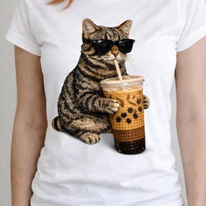 May include: White t-shirt featuring a tabby cat wearing sunglasses and drinking a bubble tea. The cat is holding the drink with both paws. The bubble tea is in a clear cup with a brown sleeve and a straw.