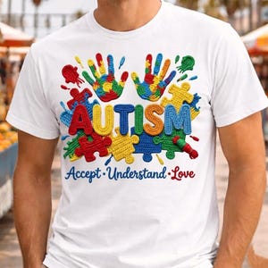 Può includere: T-shirt bianca con un design colorato. La parola "AUTISMO" è visualizzata in grandi lettere strutturate. Il design include impronte di mani e pezzi di puzzle in vari colori, con le parole "Accettare. Comprendere. Amare" sotto.