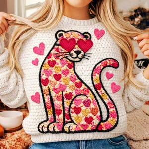 May include: Cream-colored crochet sweater featuring a large leopard design adorned with pink and gold hearts. The leopard wears heart-shaped sunglasses. Additional pink hearts are scattered across the sweater. A cozy and unique garment.