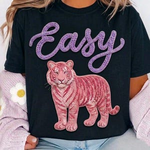 May include: Black t-shirt featuring a pink tiger design with the word "Easy" in purple, knitted-style lettering. The tiger has pink and white stripes. The shirt is a casual wear item.