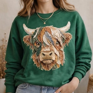 Puede incluir: Una sudadera verde oscuro con una aplicación detallada de la cara de una vaca de las Highlands. La cara de la vaca está hecha de varios parches de tela en tonos marrones, verdes y azules. La sudadera tiene cuello redondo, mangas largas y un collar dorado.