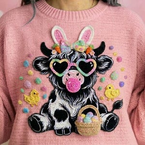 Puede incluir: Suéter rosa con un diseño de vaca en blanco y negro con gafas de sol en forma de corazón y orejas de conejo. La vaca sostiene una cesta de huevos de Pascua. El suéter tiene adornos coloridos y dos diseños de pollitos amarillos.