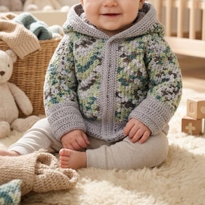 May include: A baby wearing a gray and green hooded crochet sweater with a button closure. The sweater has a camouflage-like pattern. The baby is also wearing gray pants and is sitting on a white rug.