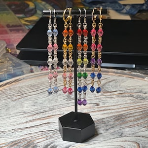 May include: A display of colorful beaded earrings. The earrings are arranged in a rainbow spectrum, with each pair featuring a different color of faceted beads. The earrings hang from a black metal stand with a hexagonal base.