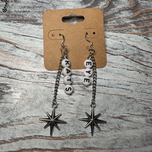 Puede incluir: Un par de pendientes colgantes con cadenas plateadas y dijes de estrella. Los pendientes tienen cuentas de letras blancas que deletrean "KATS" y "EYE". Se exhiben en una tarjeta marrón.