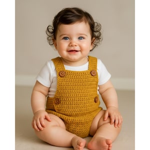Puede incluir: Un bebé sonriente con un pelele de crochet amarillo mostaza con grandes botones de madera. El pelele se lleva sobre un body blanco de manga corta. El bebé tiene el pelo rizado oscuro y ojos azules.