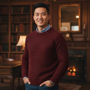 May include: A burgundy crew neck sweater with a textured knit pattern. The sweater is worn over a light blue collared shirt and paired with blue jeans. A watch with a brown leather strap is visible on the wrist.